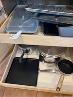 Wide angle photo of kitchen cabinet and drawer storing various pots, pans, griddle, baking sheets, cookie rack, and plastic storage containers.