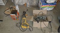 Overview of the three corded power tools showing reciprocating saw, angle grinder, and finishing sander in cardboard boxes and loose on the floor