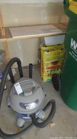 Shop Vac 6 gallon 3.0 HP vacuum with hose and accessories on a concrete floor near a green waste bin and wooden shelving.