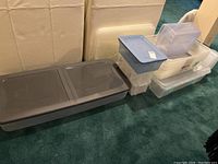 Photo showing a variety of plastic storage tubs and bins stacked against a wall, with assorted lids mostly in gray, blue, and clear plastic.