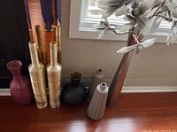 Seven vases together in a line, various shapes and colors including matte pink and black, metallic gold and brown, and ceramic ribbed vases