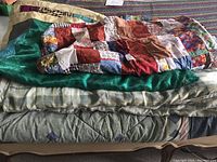 Overall view of stacked folded bedding showing comforters and quilts with a mix of solid green, striped, and patchwork colors.