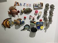 Full view of the collection showing six hand-painted fish magnets, various clip magnets, souvenir magnets, and colored round magnets attached to a white surface.