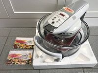 Flavorwave Turbo Oven in glass bowl with digital control lid placed in white foam packaging, with instruction manual and receipts booklet beside it.