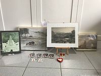 Wide view of all four framed prints displayed standing on floor along with six Snoopy memorabilia badges/buttons, heart-shaped item and wooden easel stand.