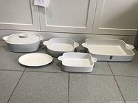 Full set laid out on floor including large and medium serving and casserole dishes, one with lid, all white ceramic with no visible chips or cracks.