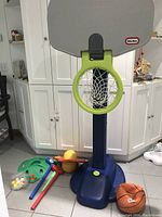 Photo of Little Tikes basketball hoop with grey backboard, green rim and net, blue adjustable base, and attached small orange basketball. Additional items visible include golf equipment and yellow jumping ball