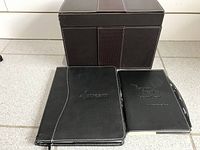 Two black leather writing portfolios with white stitching, one embossed with "Altecream", the other with "The Hamilton Collection", placed in front of a brown leather storage box with vertical stitched bands.