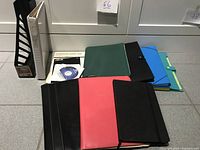 Photo displaying a collection of binders in various colors, a black leather portfolio, writing pads, and a CD in a box.