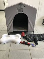 Front view of gray canvas dog house with black interior cushion. White plastic double feeder, black harness, red collar, and Kong toy in front.