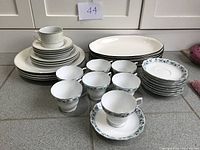 Overview of stacked dishes and cups including oval serving dishes, plates, cups and saucers on a countertop under natural light.