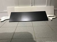 Black wooden shelf panel shown front side up on tiled floor.