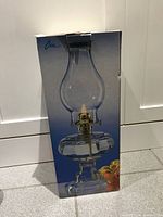 Box showing clear glass hurricane oil lamp depicted on front, lamp features classic shape with a brass burner component.