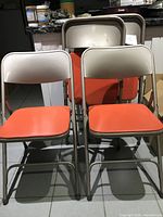 Four vintage metal folding chairs shown from front and slightly angled view, featuring red padded vinyl seats with beige metal frames.