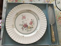 Porcelain cake plate with floral design beside the matching gold-tone handled cake server blade in original box