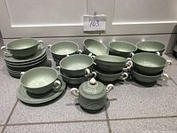 Group of 16 celadon green soup bowls with white handles, 16 matching saucers, and sugar bowl with lid on countertop