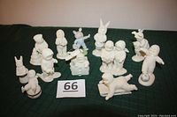 Photo showing 12 white Snowbabies figurines arranged on a dark green cloth, featuring a range of poses including sledding, standing, and some with bunny ears.