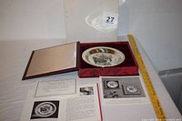 Collector plate in open presentation box showing the plate and accompanying certificates and documentation spread beside it.