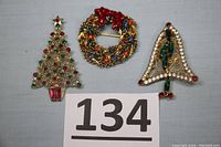 Three vintage rhinestone holiday brooches including a Christmas tree, wreath, and bell shape shown together on a gray cloth background with lot number 134.