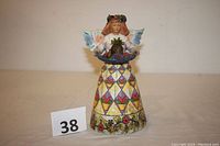 Full frontal view of Angel of Generosity figurine with a colorful skirt and floral base, holding a basket of fruit with wings visible.