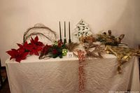 Overview of the assorted holiday decorative elements displayed on a white cloth-covered table including red poinsettias, green candles, wreath, ceramic Christmas tree, ribbons, and berry sprays.
