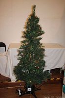 Photo of a 6-foot artificial Christmas tree with fiber-optic multicolor lights and additional string lights, displayed indoors against a neutral background, showing the full height and black plastic base.