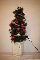 Full view of the green artificial Christmas tree decorated with multicolor lights, pink ribbon, red and blue ornaments, on a white distressed wooden stand.