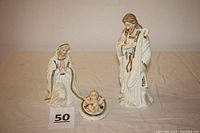Wide photo showing all three figurines of the Holy Family in white with gold detailing: standing Joseph, kneeling Mary, and Infant Jesus in a small oval bowl/manger.
