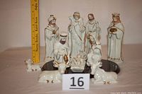 Photo shows nine ivory ceramic nativity figurines with gold trim arranged on a wooden base, including human characters and animals; a ruler in background indicates tallest is around 7.5 inches tall.