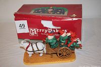Full view of the porcelain figurine set showing the three carolers on the cart pulled by the white goat with a yellow base and original red Merry Makers box in background.
