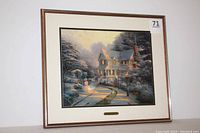 Framed print showing winter night scene with house, snowman, snow-covered trees and bushes, glowing street lamp, and pathway covered with snow.