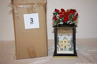 Front view of the Thomas Kinkade inspired sparkling lantern showing the winter house scene inside and festive greenery decoration on the top, next to its cardboard box.