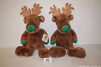 Two brown plush stuffed reindeer toys with green noses, red and green mittens, and brown antlers sitting side-by-side.
