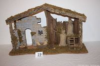 Front view of the large rustic manger showing moss roof, stone wall with window and doorway, wooden planks on side with hanging figures and ladder.