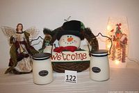 Front view of two Milkhouse Candle Co. candles, snowman welcome wreath, angel figurine, and lit vase.