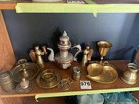 Shelf display of copper teapot with dragon motif center surrounded by brass angels holding candle holders, brass goblet, candlesticks and small decorative dishes.