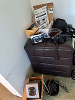 Vintage cameras and accessories arranged on a brown ottoman: Kodak Instamatic X30 camera, Pentax camera, Canon camera, flash unit box, and some cables.