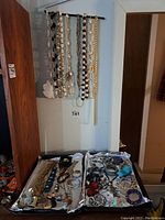 Photo of multiple necklaces hanging on black rod mounted on wall and various bracelets and other jewelry laid out below on trays.