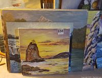 Stack of landscape paintings including a sunset rocky shore scene showing signature and lot number 858