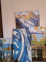Four oil on canvas paintings stacked and leaning against a wall. Mountain, abstract blue design, lake landscape, and tree-focused landscape.