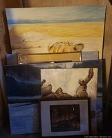 Photo showing five paintings stacked, including framed artwork and an unframed canvas with a cheetah in the background. One framed piece has cracked glass visible.