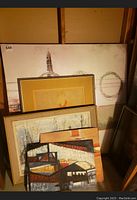 Stack of five framed abstract paintings. Largest visible painting about 47x47 inches with abstract circular and figure motifs in beige, brown, and orange hues. Smaller paintings with varied abstract themes and frames leaning against wall.