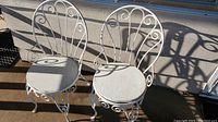 Two vintage white wrought iron patio chairs with round wooden seats on an outdoor carpeted surface under sunlight.