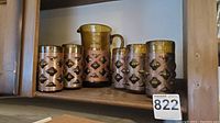 Amber glass pitcher and 5 glasses with copper diamond patterned copper bands on a wooden shelf