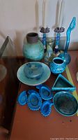 Photo showing a variety of blue and turquoise glass and pottery vases, a plate, two tall candlesticks with floral details, a small blue vase, triangular dish, and decorative blue glass pieces on a surface.