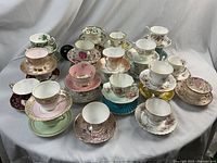 Overall view of 29 assorted tea cups and saucers on white draped background with green apple reference