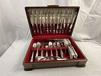 Wooden flatware storage case open showing silver-plated flatware arranged in red felt interior. Contains top row of dinner knives and bottom tray with assorted spoons, forks and serving utensils.