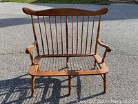 Front view of a wooden spindle back bench frame showing vertical spindles and armrests on an asphalt background.