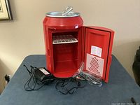 Red Coca Cola branded mini thermal electric cooler/warmer with door open showing interior shelf and included power adapters on table.