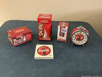 Full view of all Coca-Cola collectibles included in the lot arranged on a table: alarm clock, stone coasters, salt and pepper set, disposable coasters tin, and jigsaw puzzle box.